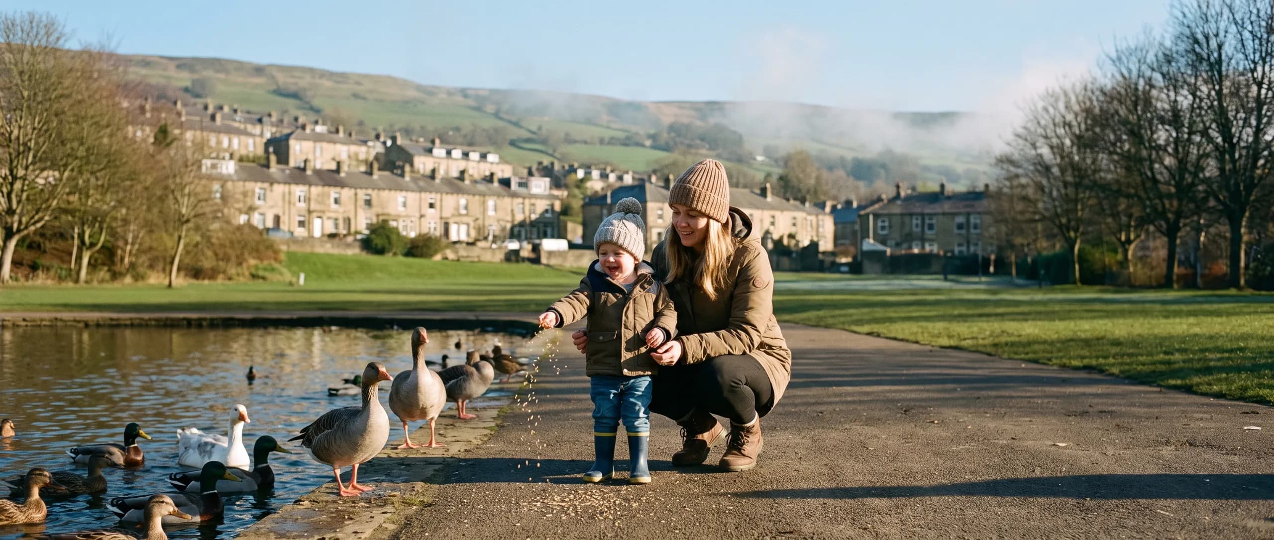 A mum and her toddler feeding the ducks at a park pond in a Welsh valley town, terraced houses on the hillside behind.