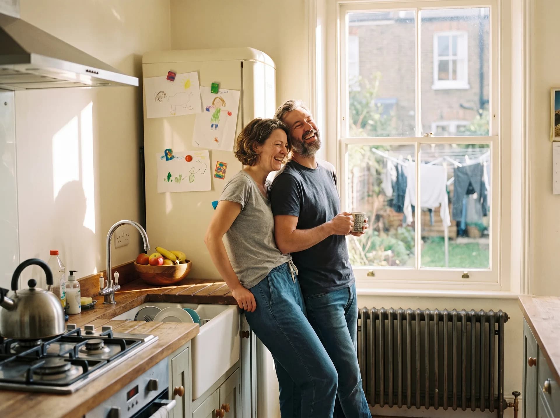 A couple in their forties laughing together over mugs of tea by the window of their terraced-house kitchen on a sunny morning.